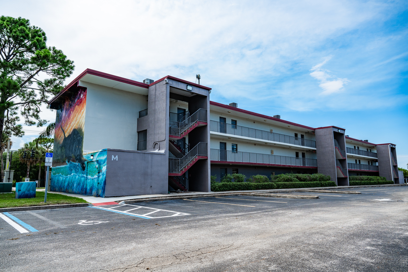 A three story apartment building with covered stairwells facing a parking lot. A large colorful mural covers one end of the building